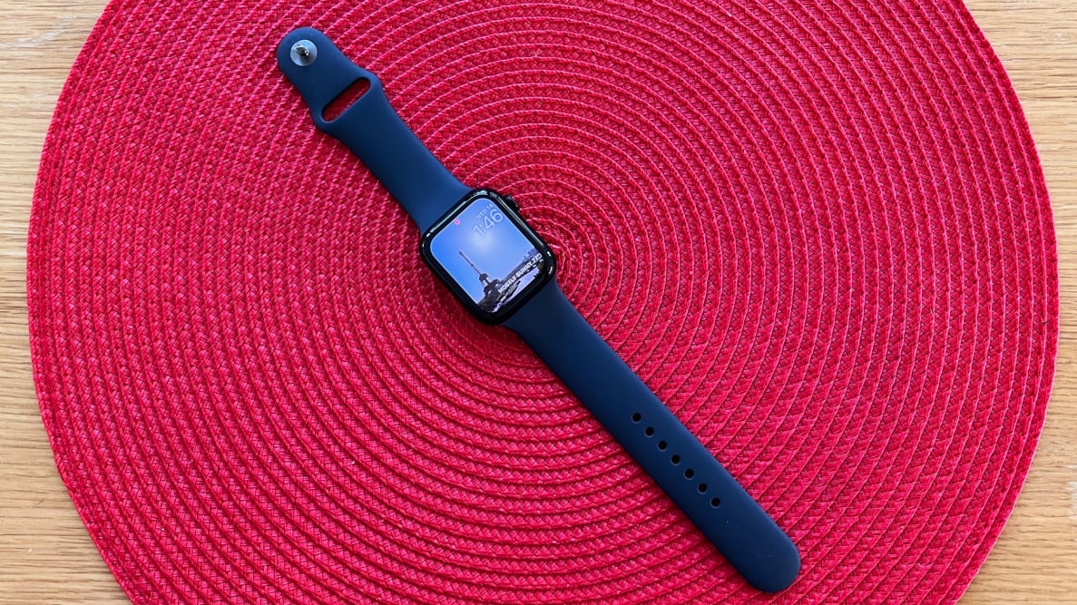 The Apple Watch SE with black band sits on a circular red background on a table.