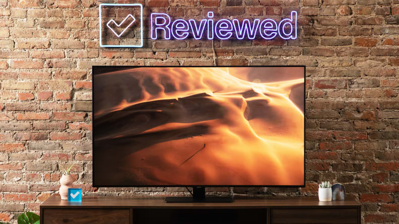 Samsung Q80B TV in front of brick wall under neon sign indoors with sand dunes on screen.
