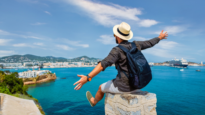 A traveler wearing a hat and backpack sits on a stone ledge, arms outstretched, overlooking a scenic coastal city.