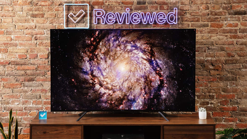 The Hisense U7H displaying an image of a galaxy, sitting on a wooden table in front of a brick wall that has a neon sign that says Reviewed.