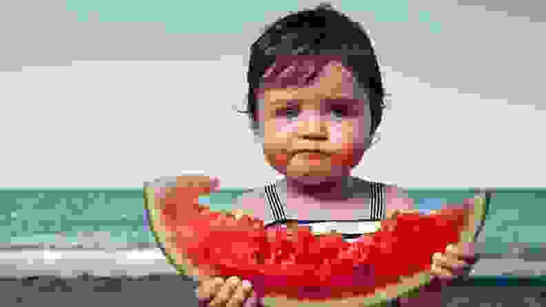A baby eating a watermelon on the beach.