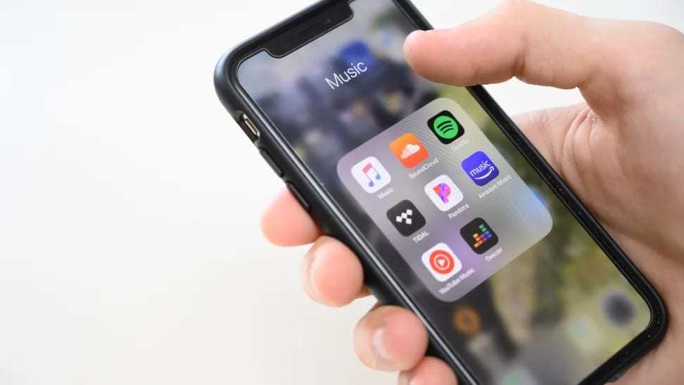 An iPhone X being held, with a folder of music apps shown on-screen. The apps include Apple Music, Soundcloud, Spotify, Tidal, and Pandora.