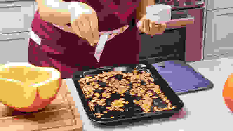 A person holding a white bowl, seasoning the naked pumpkin seeds on the metal sheet.