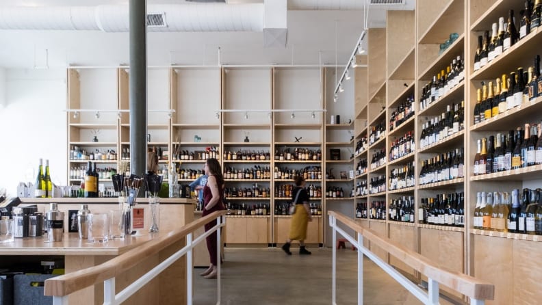 Four people shop for wine in a brightly lit natural wine store with light wood shelves filled with colorful bottles.