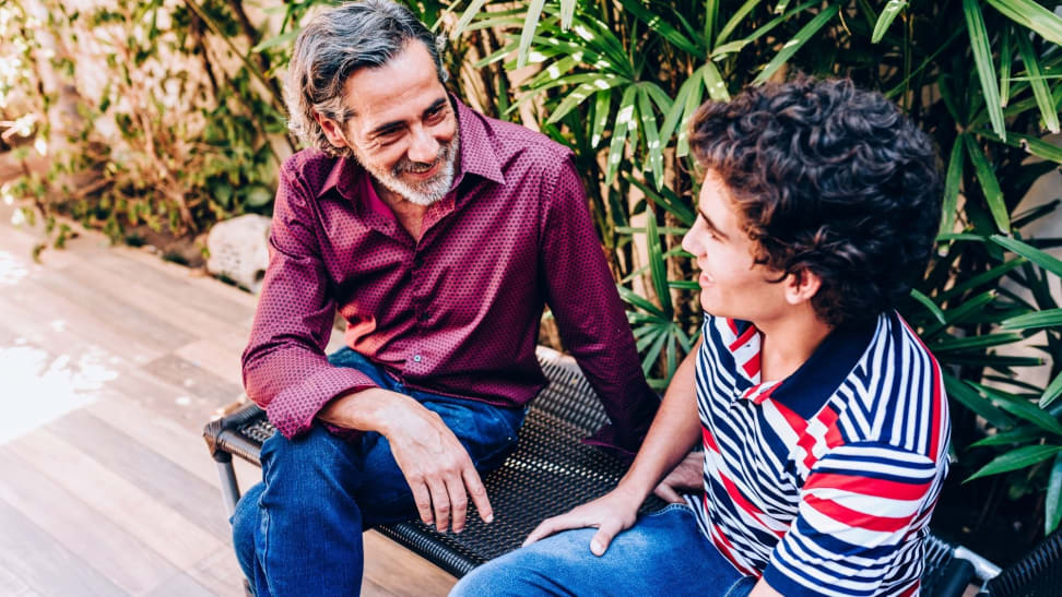 A dad talks to his teenage son as they sit on an outdoor bench.