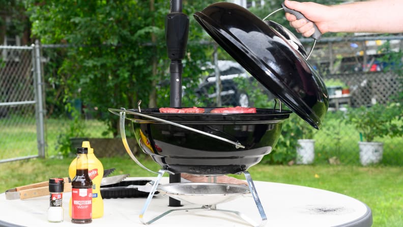 A hand pulling the lid off of a small charcoal grill