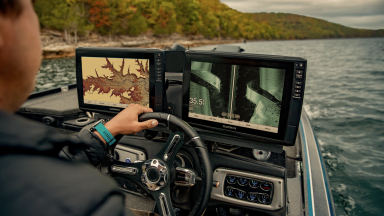 A fishing enthusiast is seated at the helm of a boat, one hand on the wheel. Colorful autumn trees are visible in the distance. A Garmin fish-finder display is mounted at the helm.