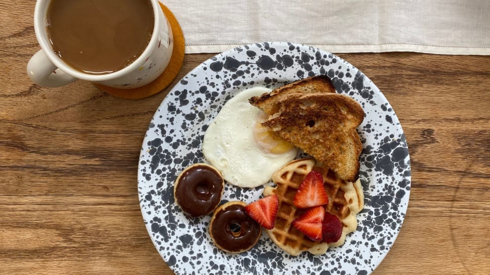 Mini toast, donuts, waffles, and a fried egg on a white and grey speckled plate accompanied by a mug of hot coffee.