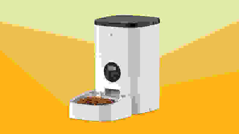 A white automatic pet feeder with food in bowl against a varied yellow background.