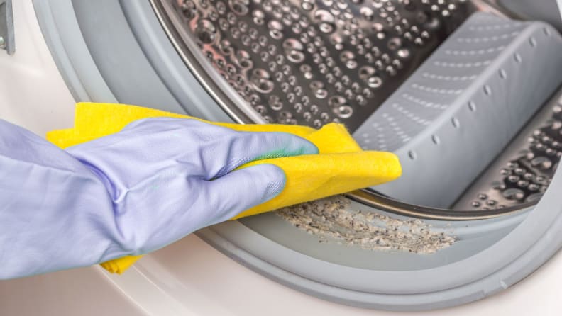 Una mano con un guante de goma se estira hacia el marco y pasa una esponja por la junta sucia de una lavadora.