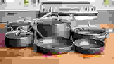 A collection of steel grey All-Clad pots and pans on a kitchen island in the foreground, a stovetop and counter in the background.