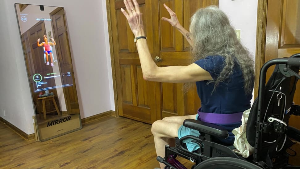 Person sitting in wheelchair in front of the lululemon Studio mirror with arm raised above head during guided fitness class.