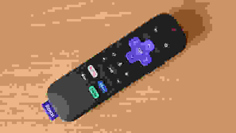 Roku Express 4K+ remote is all black with colorful buttons on top, including quick keys for streaming services like Netflix, Disney+, Hulu, and Apple TV+. It sits atop a yellow colored woodgrain table.