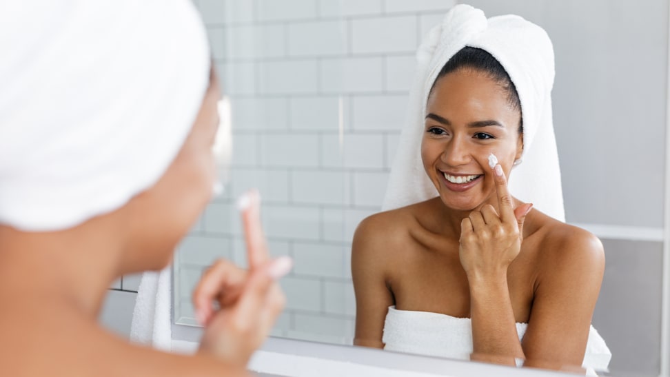 A person standing in front of a large bathroom mirror applying a moisturizer to her face while wearing a towel around her body and hair.