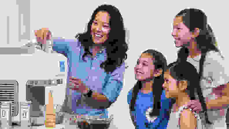 A parent and several children gather, smiling, around a large white machine. Ice-cream cones and toppings are on the table beside it.