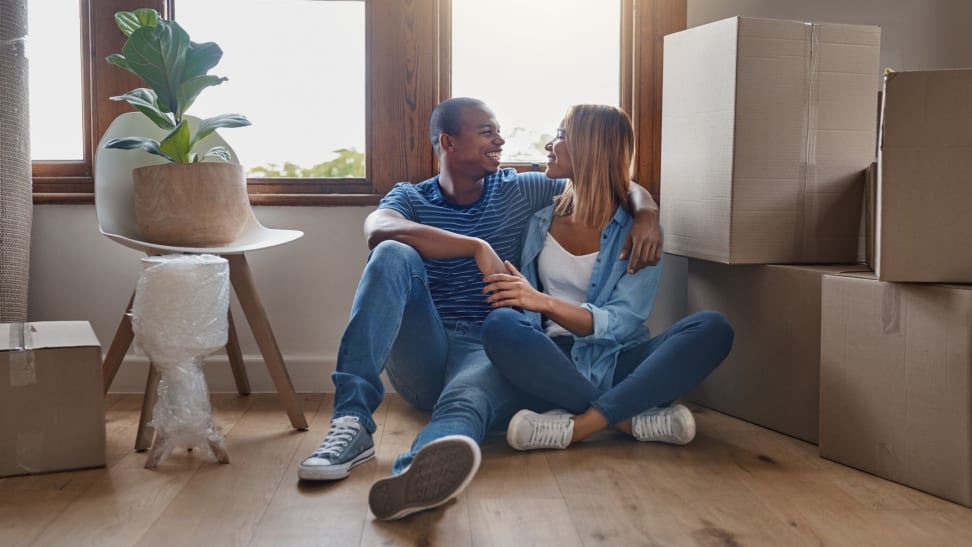 An unmarried couple sits on the floor of an unfurnished house with moving boxes piled beside them