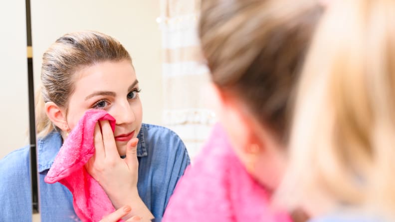 A woman stares into a large mirror and wipes her skin with a hot pink Makeup Eraser towel.