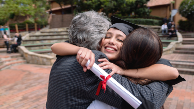 Girl smiling wearing cap and gown hugs parents while holding a diploma in hand.