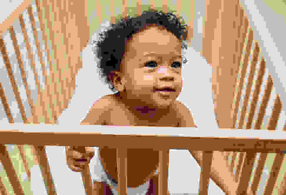 A small child stands up in a wooden crib.