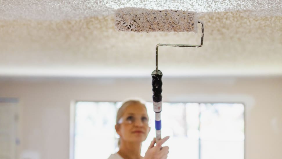 Blonde woman wearing safety glasses and painting the ceiling white with a paint roller.