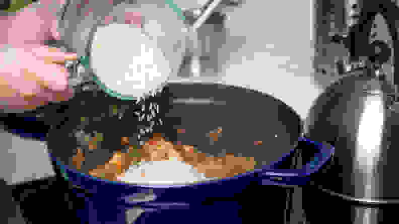 A bowl of jasmine rice being poured into a big pot of jambalaya cooking on a stove.
