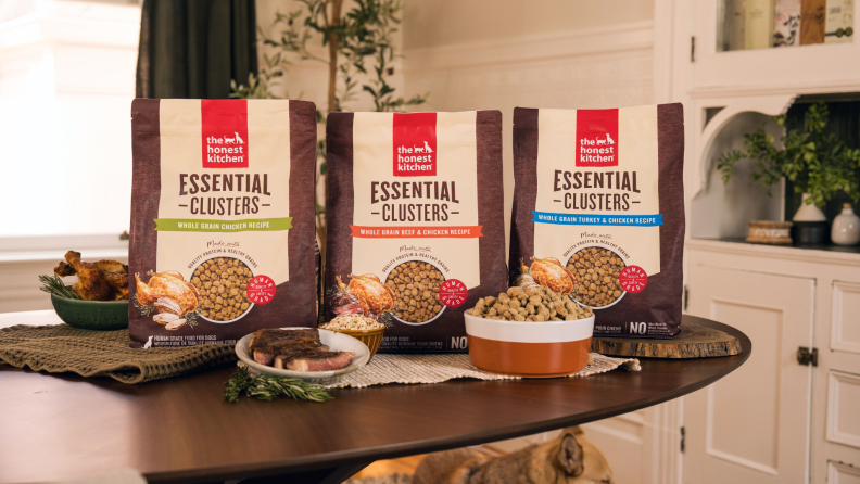 Three bags of The Honest Kitchen dog food Essential Clusters in chicken, beef & chicken, and turkey & chicken recipes displayed on a table with fresh ingredients.