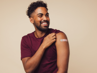 Man against brown background smiling after receiving vaccination, showing his arm with a bandage at point of injection