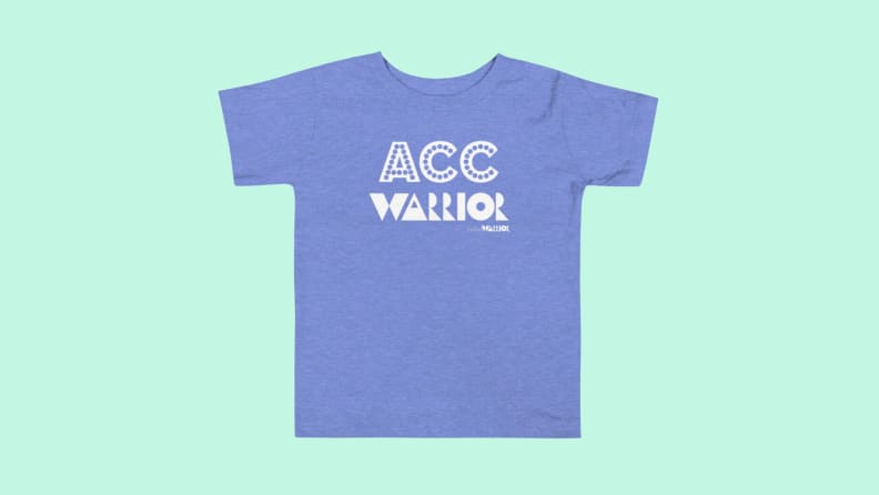 A blue Littlest Warrior T-shirt on a green background.