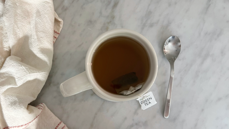 A mug of green tea, a spoon, and a dish cloth on a marble counter.
