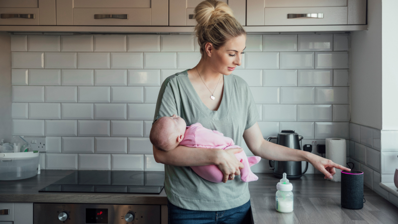 Person holding baby in kitchen while engaging with smart speaker.