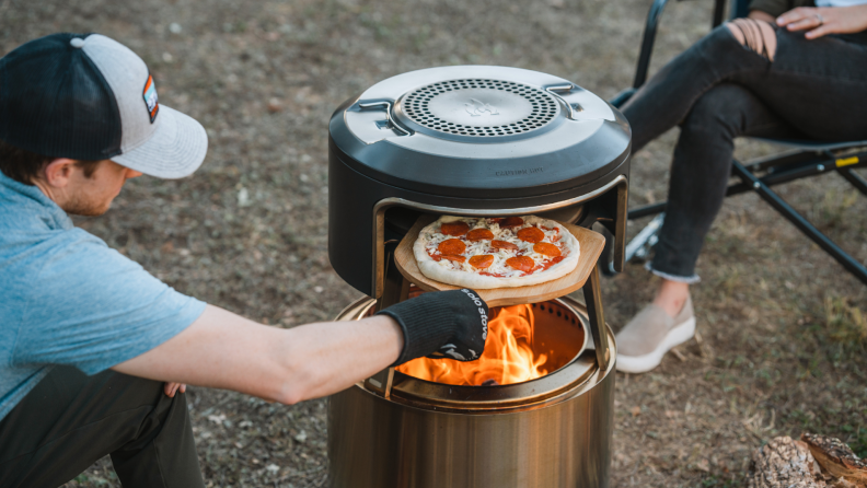 Person wearing glove uses wooden pizza peel to insert uncooked pizza inside of Solo Stove Pi Fire pizza oven with open flame.