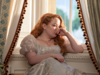 An image of Penelope Featherington in the Netflix series "Bridgerton." Penelope sits in a window seat, gazing wistfully outward.