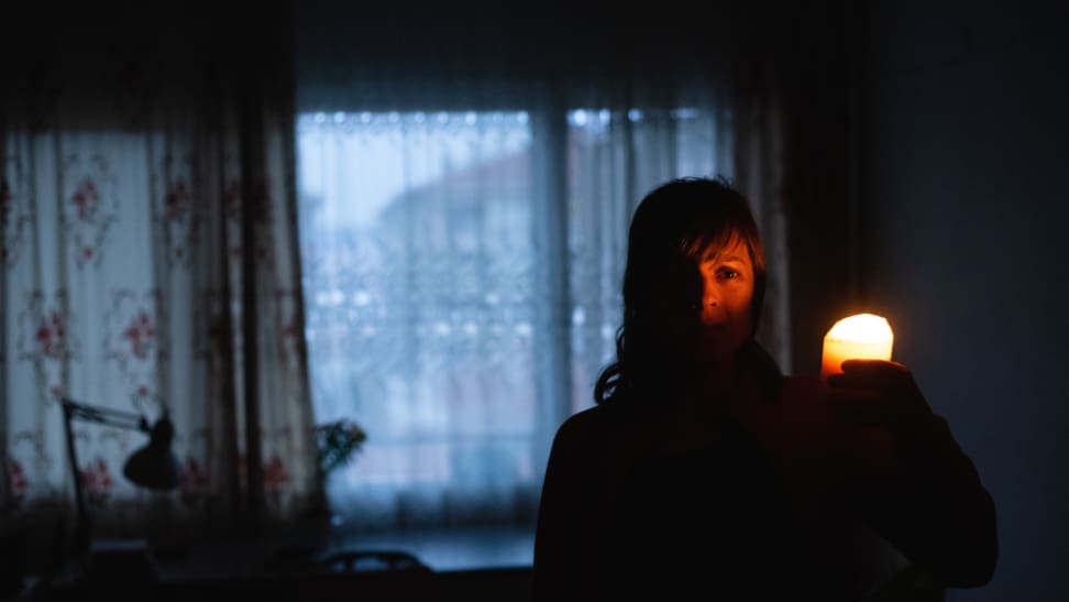 一个人站在黑暗的房间里，身后有一扇窗户，拿着一支点燃的蜡烛对着脸
