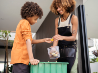 A brother and sister of color add food scraps to a large green compost bin