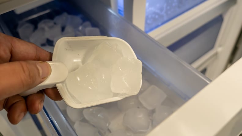 One hand reaches into the frame with a small scoop, removing some ice cubes from the ice maker.