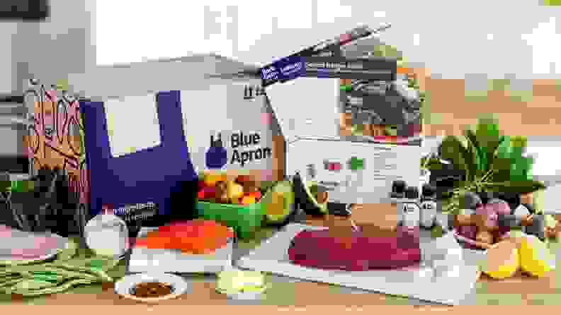 A Blue Apron box with surrounding ingredients, including produce and fresh fish, sitting on a countertop