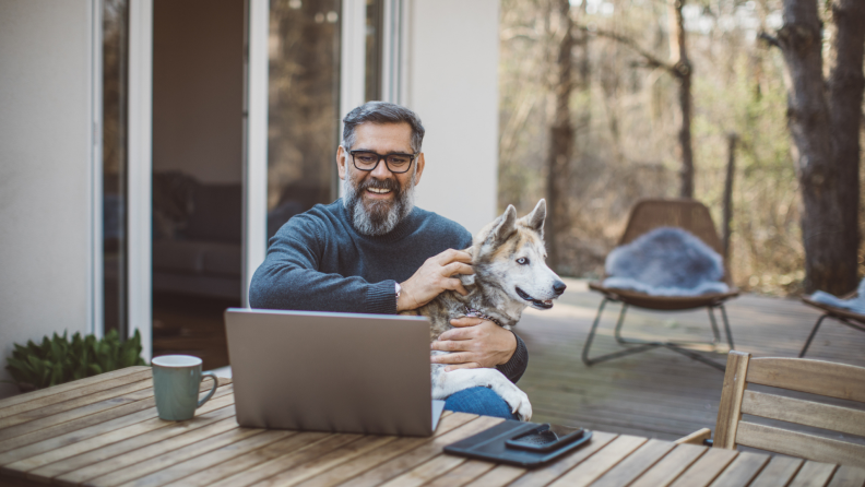 Man sitting down at table outside while smiling at computer screen and holding a husky dog.