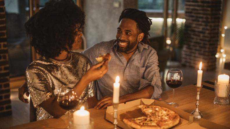 A smiling couple shares a pizza and wine by candlelight.