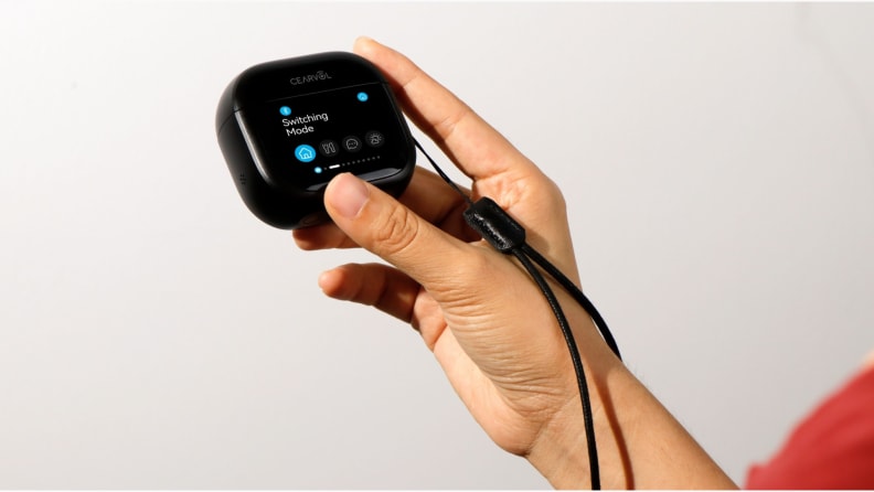 A hand holds a compact charging case with a touchscreen display for OTC hearing aids, showing settings being adjusted against a plain background.