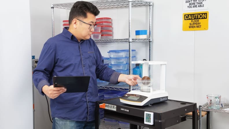 Lab technician placing plastic Tupperware container with floral foam ball inside on scale.