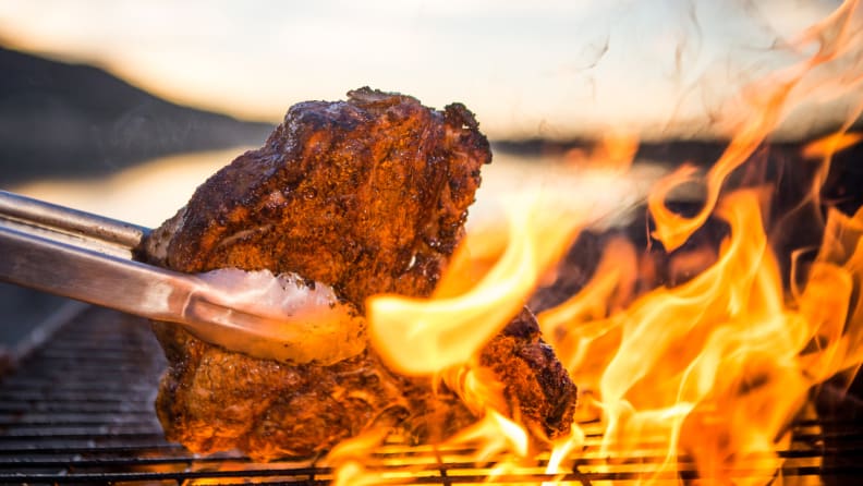 Grill tongs flipping a piece of meat over a flame-covered grill