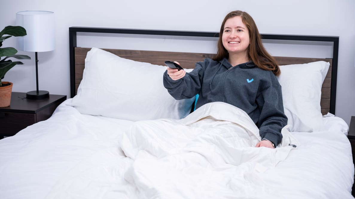 writer Sara Tabin sits against a headboard with the Baloo blanket draped over her legs. She is smiling and using a remote, staring into the distance.