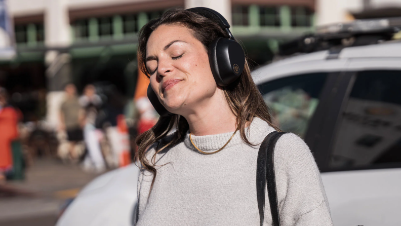 A woman standing outdoors with her eyes closed, enjoying music through sleek over-ear headphones, basking in the sunlight with a serene expression.
