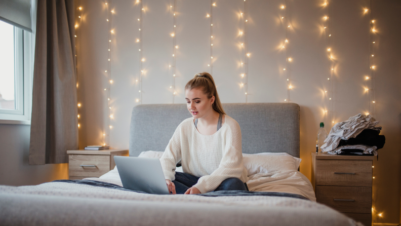 A girl sitting on her bed with a curtain of fairy lights behind her.