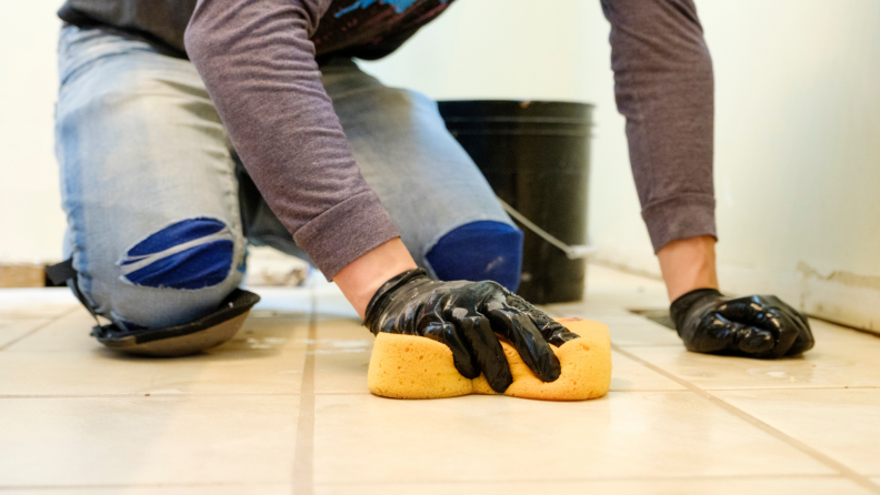 Person on hands and knees using sponge on tiles to clean up grout.