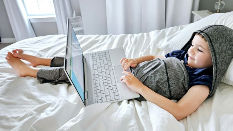 A boy sits on a bed looking at his laptop screen while wearing a gray Hug Sleep pod.