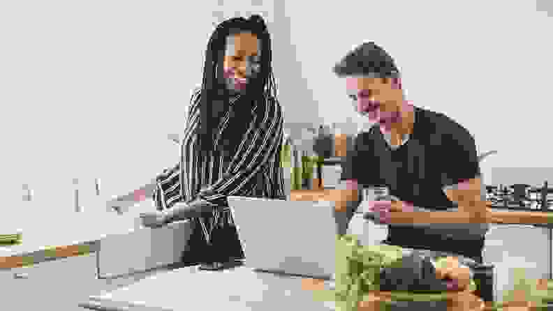 A person standing at the kitchen sink looking at their partner who is seated at an island with a computer