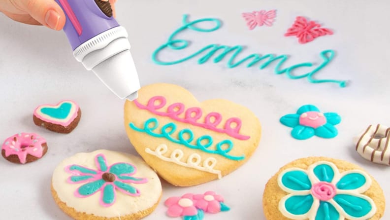 Person using pen to decorate heart-shaped cookie surrounded with baked goods.