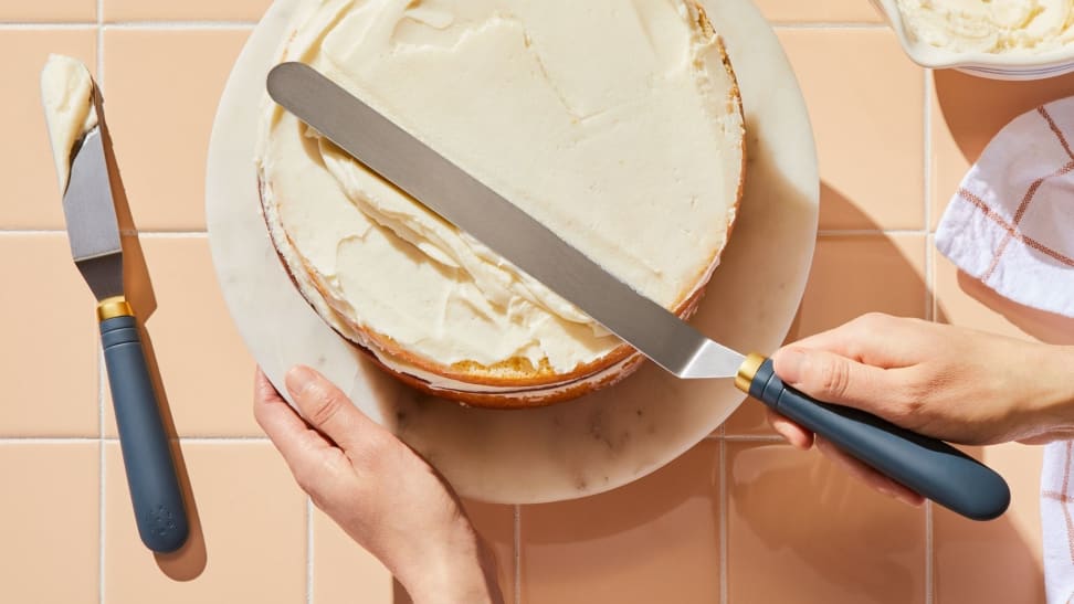 One hand holds a cake stand steady while the other spreads vanilla frosting across the cake using a large offset spatula.