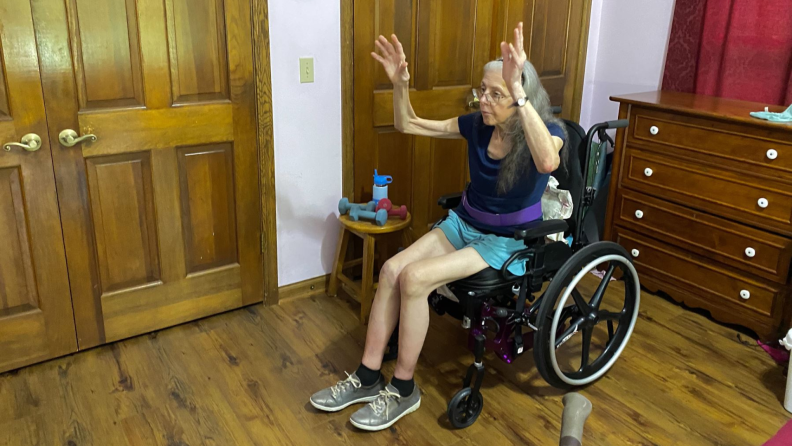 Person sitting in wheelchair in front of the lululemon Studio mirror with arm raised above head during guided fitness class.